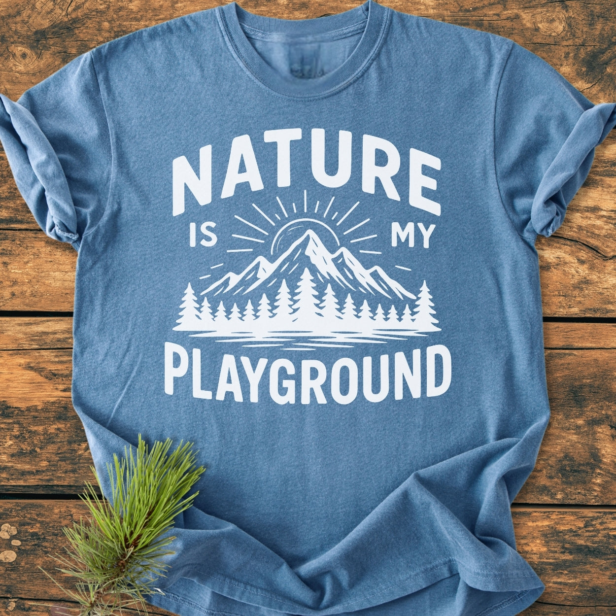Nature Is My Playground - Vintage Wash Tee features a white graphic of a sun over mountains and trees on a blue T-shirt, styled with a pine branch, highlighting its relaxed fit.