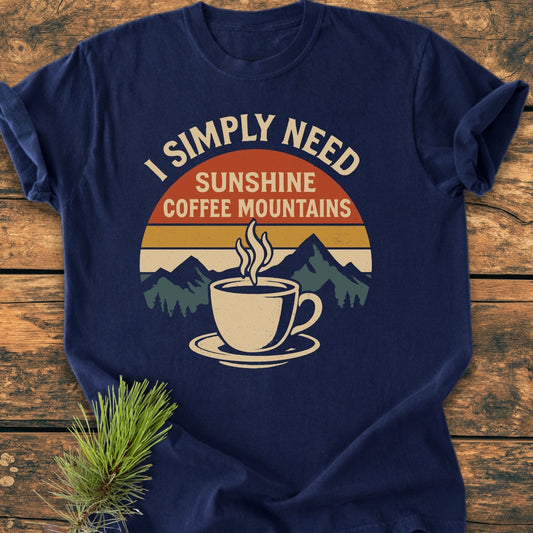 Retro Sunrise Coffee & Mountains - Vintage Wash Tee features a circular retro graphic with sunset stripes and mountains, topped by a steaming coffee cup. Laid on wood, adjacent to pine greenery.