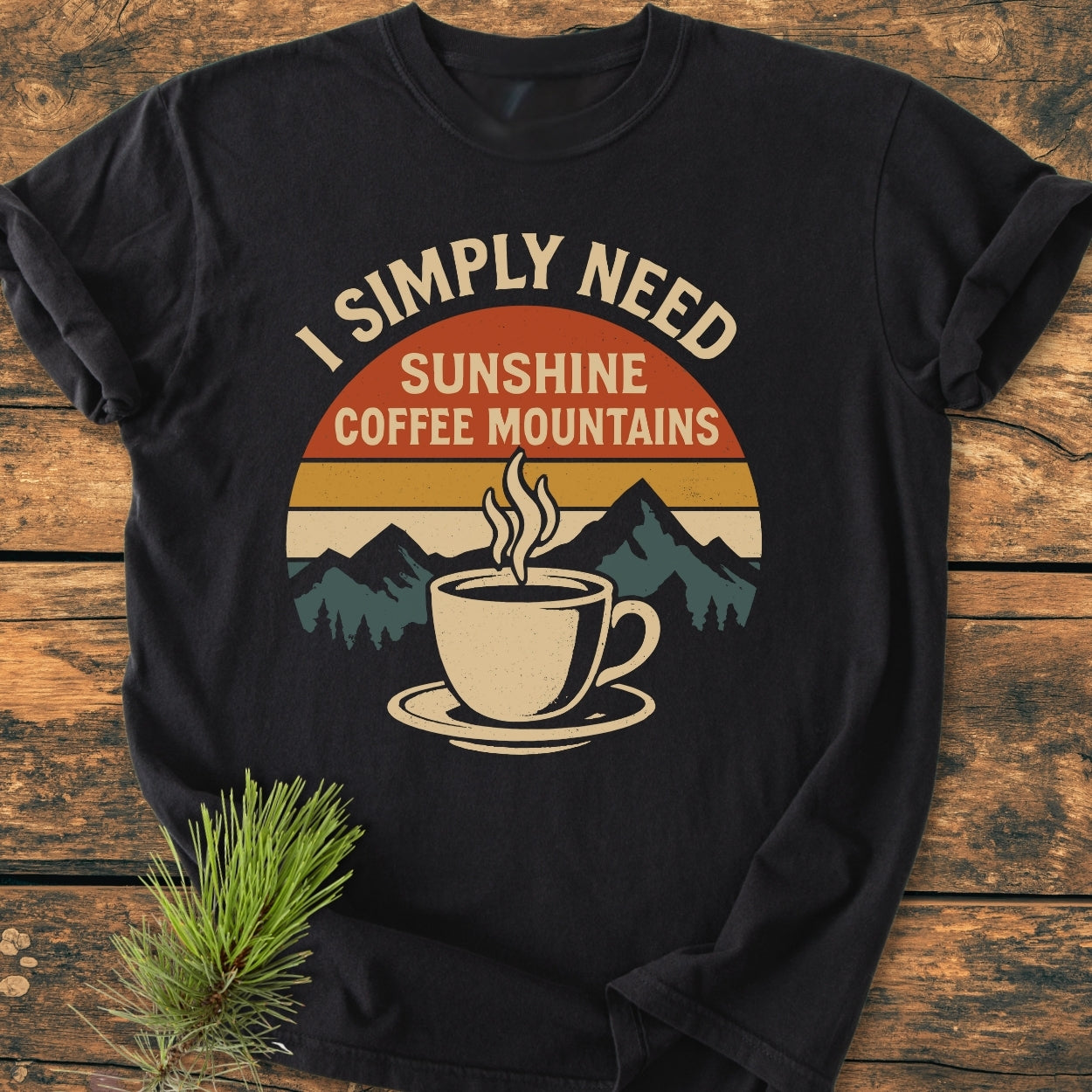 Retro Sunrise Coffee & Mountains - Vintage Wash Tee displayed on wood, features a retro sunset graphic with teal mountains, a steaming coffee cup, and the slogan “I SIMPLY NEED SUNSHINE COFFEE MOUNTAINS.”