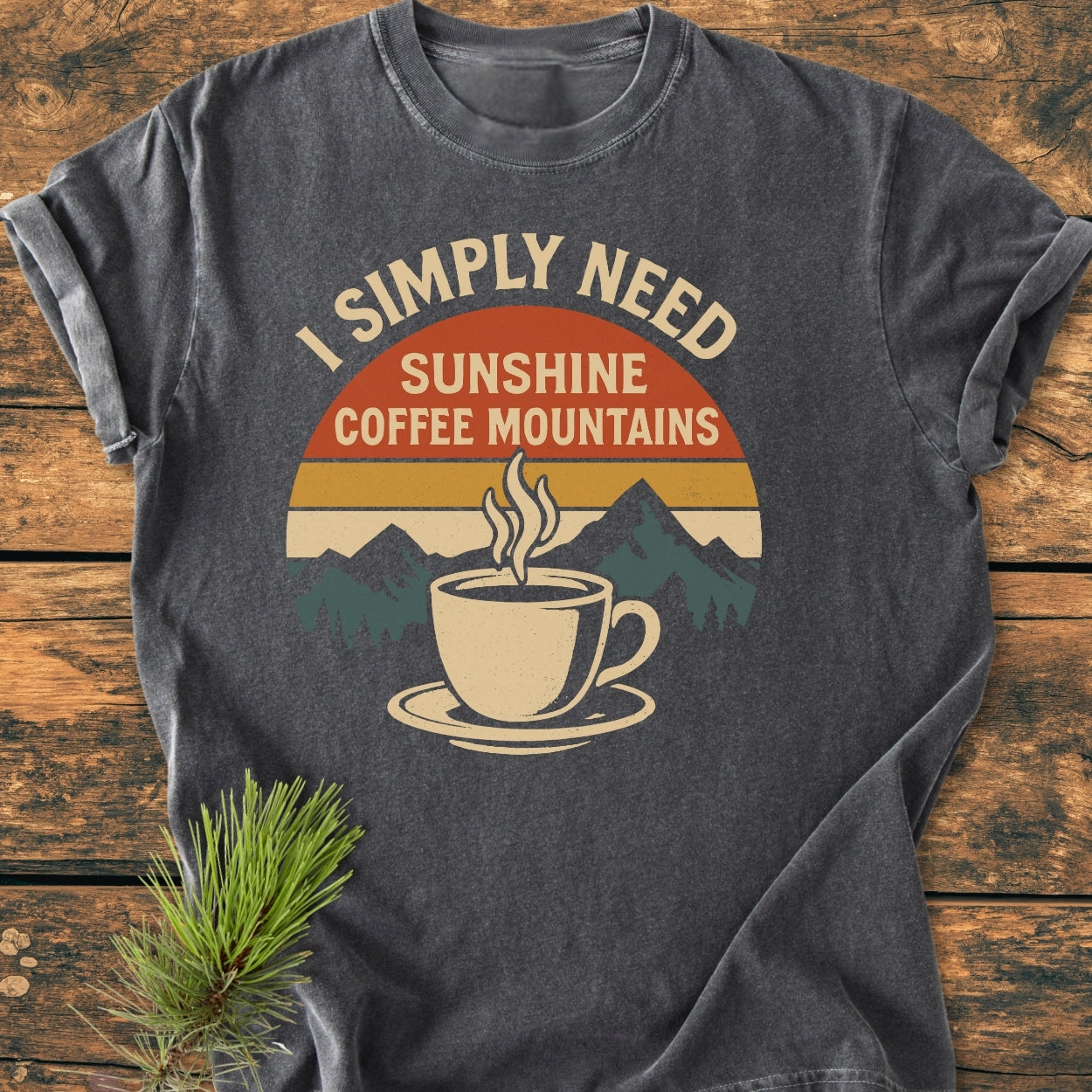 Retro Sunrise Coffee & Mountains - Vintage Wash Tee displayed on wood, features retro graphic with text “I SIMPLY NEED”, colorful stripes, mountain silhouette, coffee cup, and pine sprig beside it.