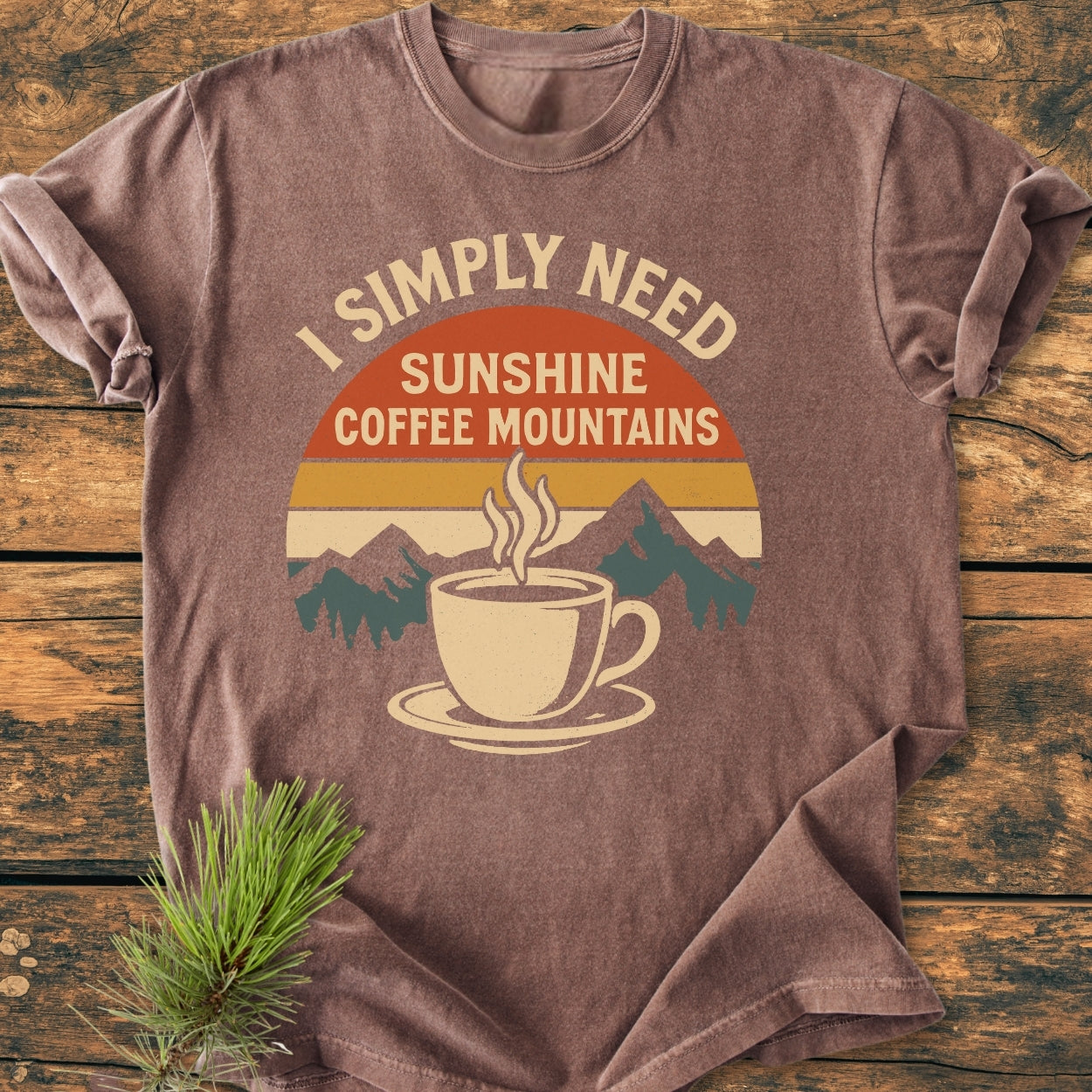 Retro Sunrise Coffee & Mountains - Vintage Wash Tee featuring a retro graphic of a sunrise, coffee cup, and mountains, laid on rustic wood with a pine sprig, highlighting its soft, relaxed fit.