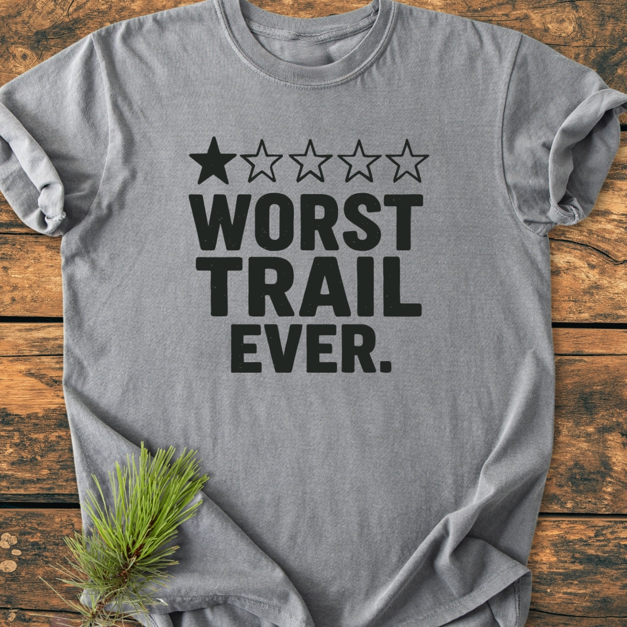 Heather-gray Worst Trail Ever - Vintage Wash Tee with a one-star rating graphic, laid on wood. Features bold black text and a pine sprig accent, showcasing a casual, relaxed fit.