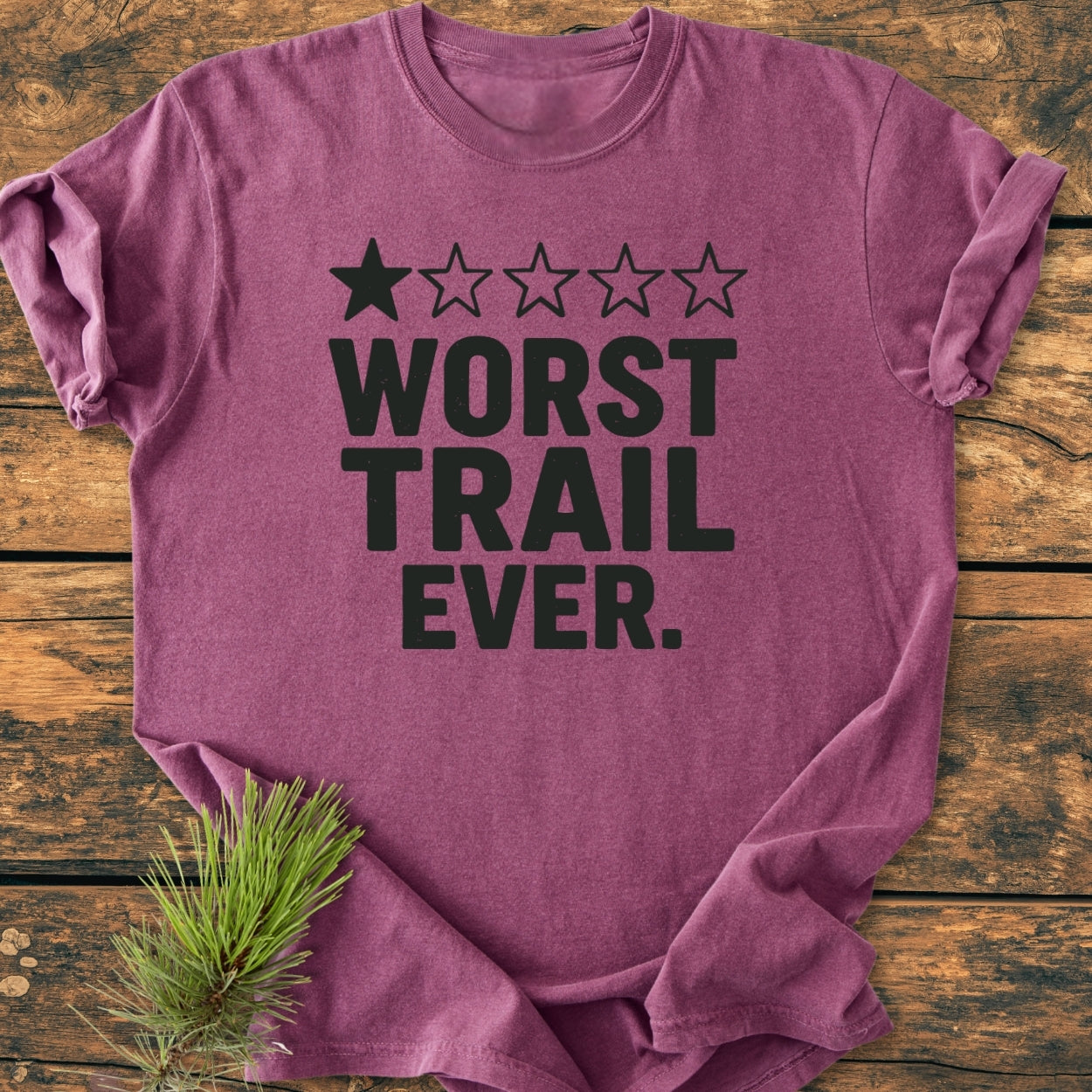 Worst Trail Ever - Vintage Wash Tee, maroon crew-neck with bold black one-star rating and slogan, laid flat on wood. Features pine needle accent, soft-washed 100% ring-spun cotton for comfort and durability.