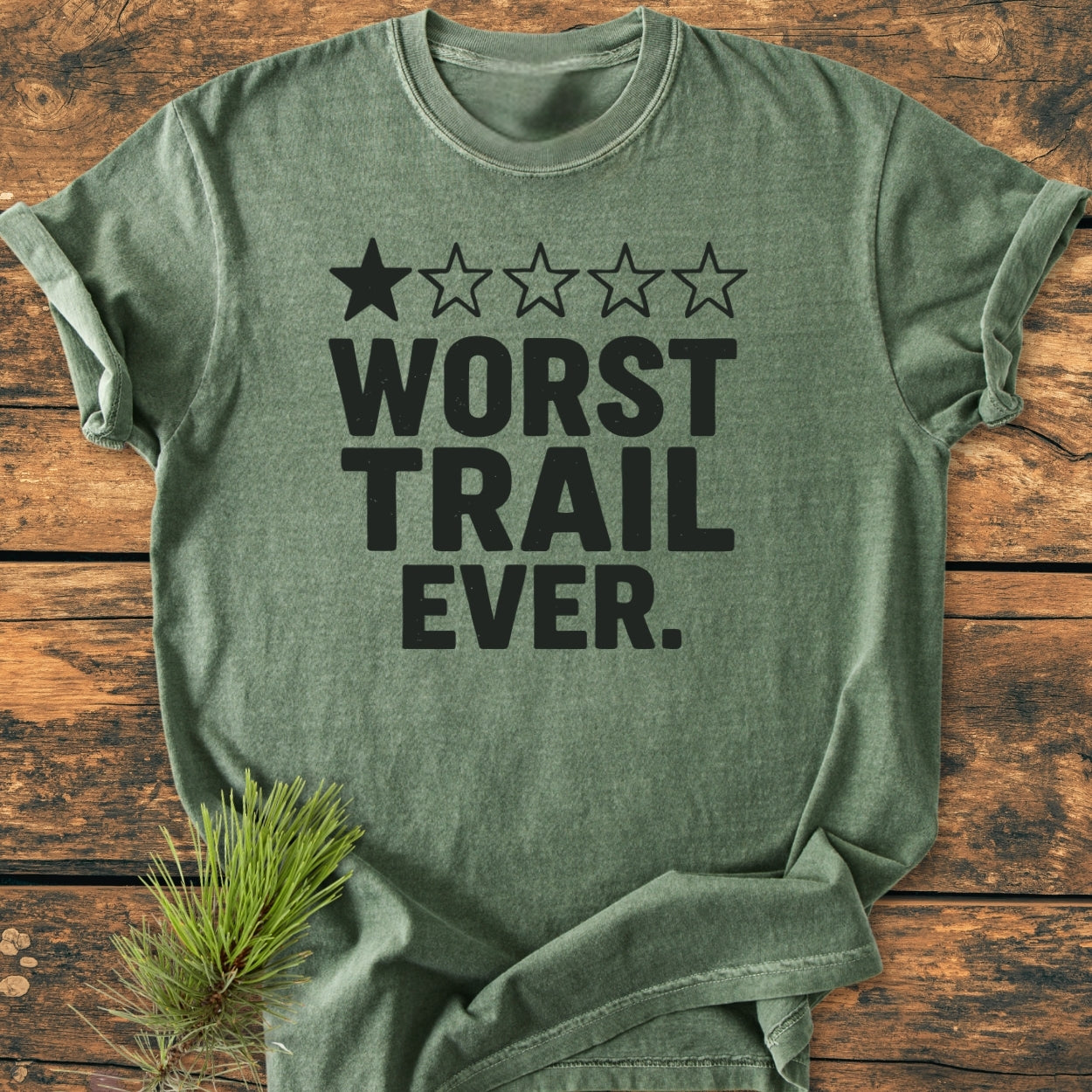 Worst Trail Ever - Vintage Wash Tee showcasing a forest-green shirt with a bold slogan and one-star rating, laid flat on wood, complemented by a pine sprig, highlighting its relaxed, cozy fit.