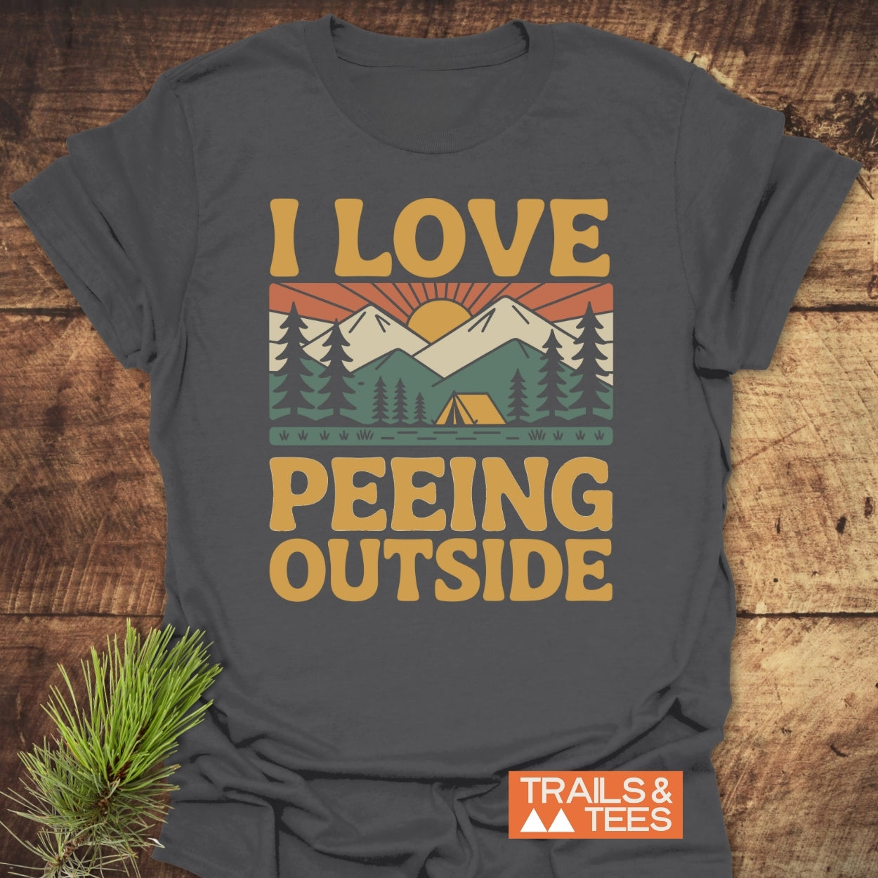 I Love Peeing Outside T-Shirt displays a retro outdoor scene with trees, tent, mountains, and sun, plus “TRAILS & TEES” logo, designed for comfort and adventure in soft ring-spun cotton.