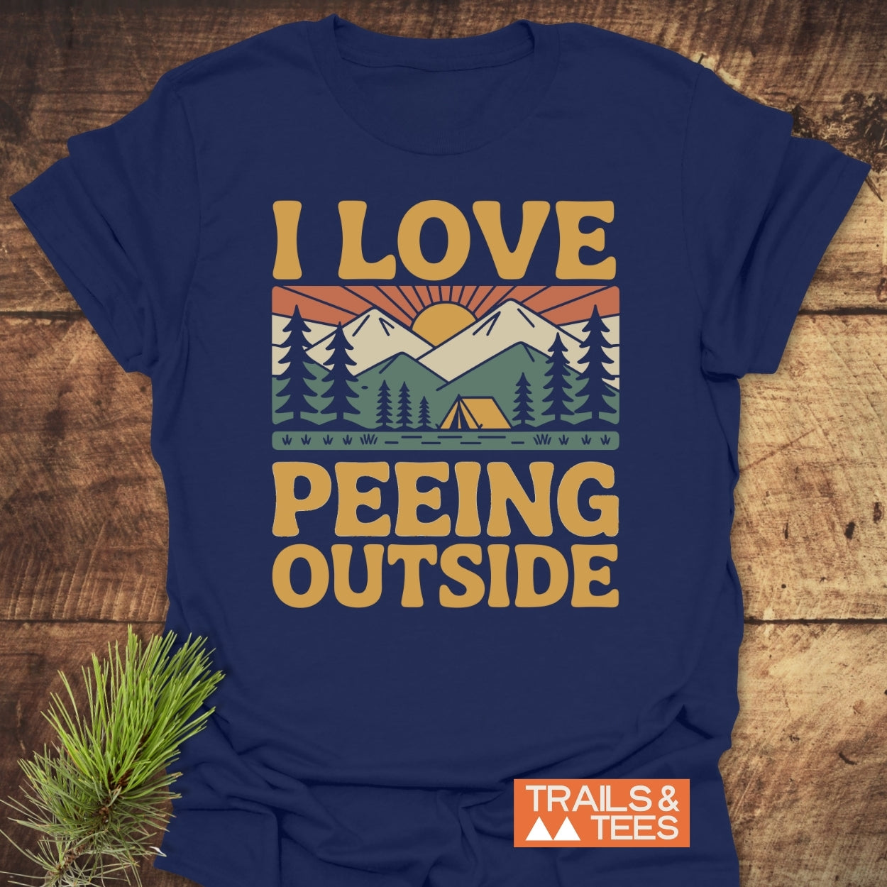 I Love Peeing Outside T-Shirt featuring an outdoor scene with pine trees, a tent, mountains, and rising sun. 'TRAILS & TEES' logo in corner, designed for comfort and outdoor enthusiasts.