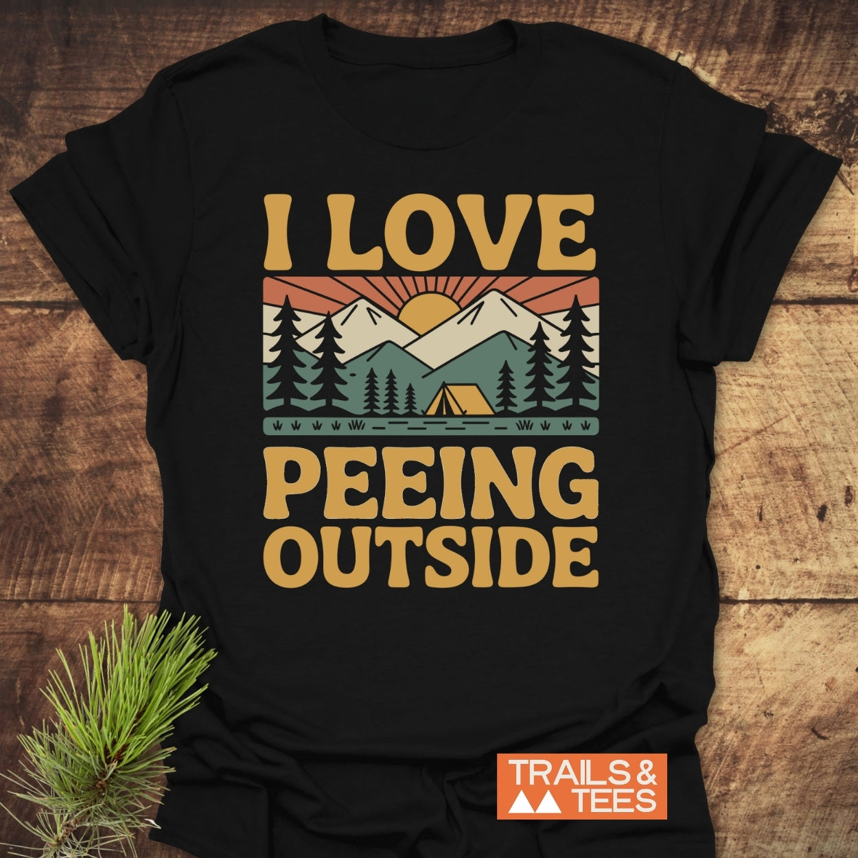 I Love Peeing Outside T-Shirt on wooden surface, featuring a retro campsite graphic with mountains and a sun. Includes durable prints, taped shoulders, and Trails & Tees logo, embodying outdoor adventure spirit.