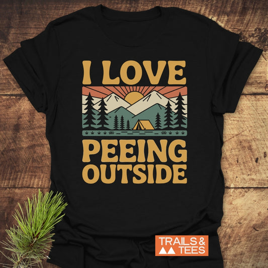 I Love Peeing Outside T-Shirt on wooden surface, featuring a retro campsite graphic with mountains and a sun. Includes durable prints, taped shoulders, and Trails & Tees logo, embodying outdoor adventure spirit.
