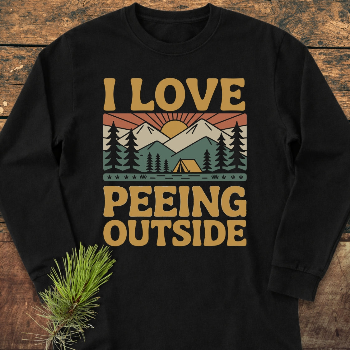 I Love Peeing Outside Long-Sleeve Tee featuring a retro camping graphic with mountain peaks, tent, and fir trees. Laid flat with a pine branch, highlighting its humorous outdoor theme and comfortable, relaxed fit.