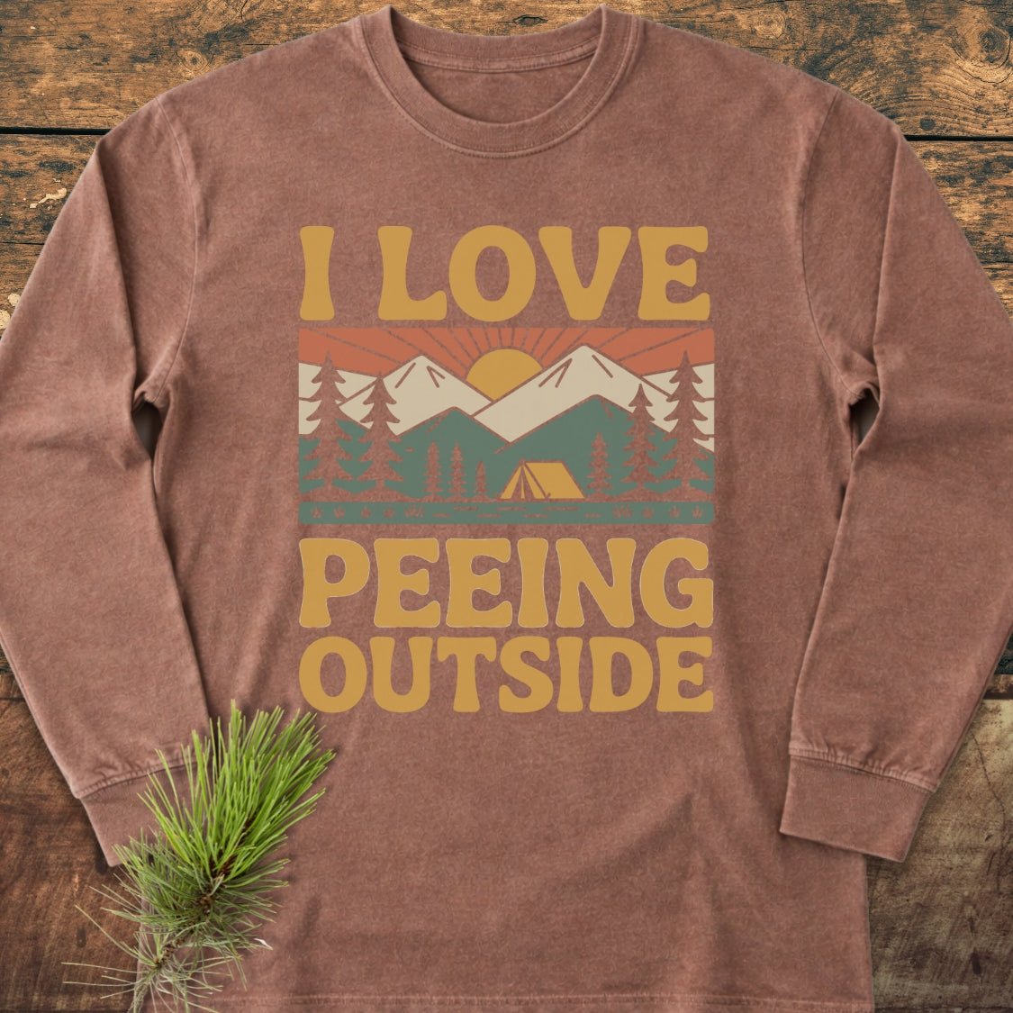 I Love Peeing Outside Long-Sleeve Tee featuring a retro campsite graphic with mountains and trees on a brown backdrop, laid on wood with a pine branch beside it. Made of soft, 100% ring-spun cotton.