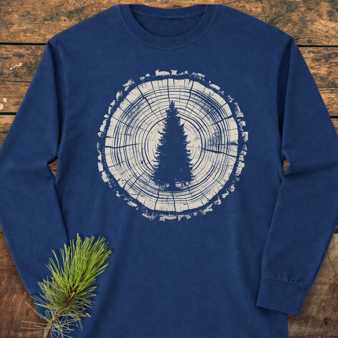 Rooted In The Wild Long-Sleeve Tee featuring a white stylized tree-ring design with a central evergreen silhouette, displayed on a rustic wood surface, alongside a pine branch. Made from 100% ring-spun cotton.