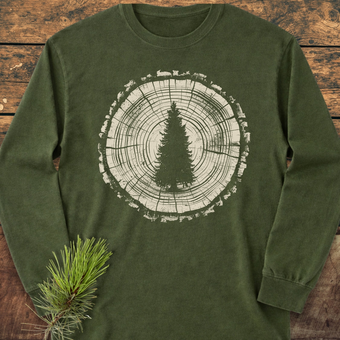 Rooted In The Wild Long-Sleeve Tee features a forest-green design with a white tree trunk graphic and pine silhouette, laid on wood, styled with a pine branch, emphasizing its natural theme.