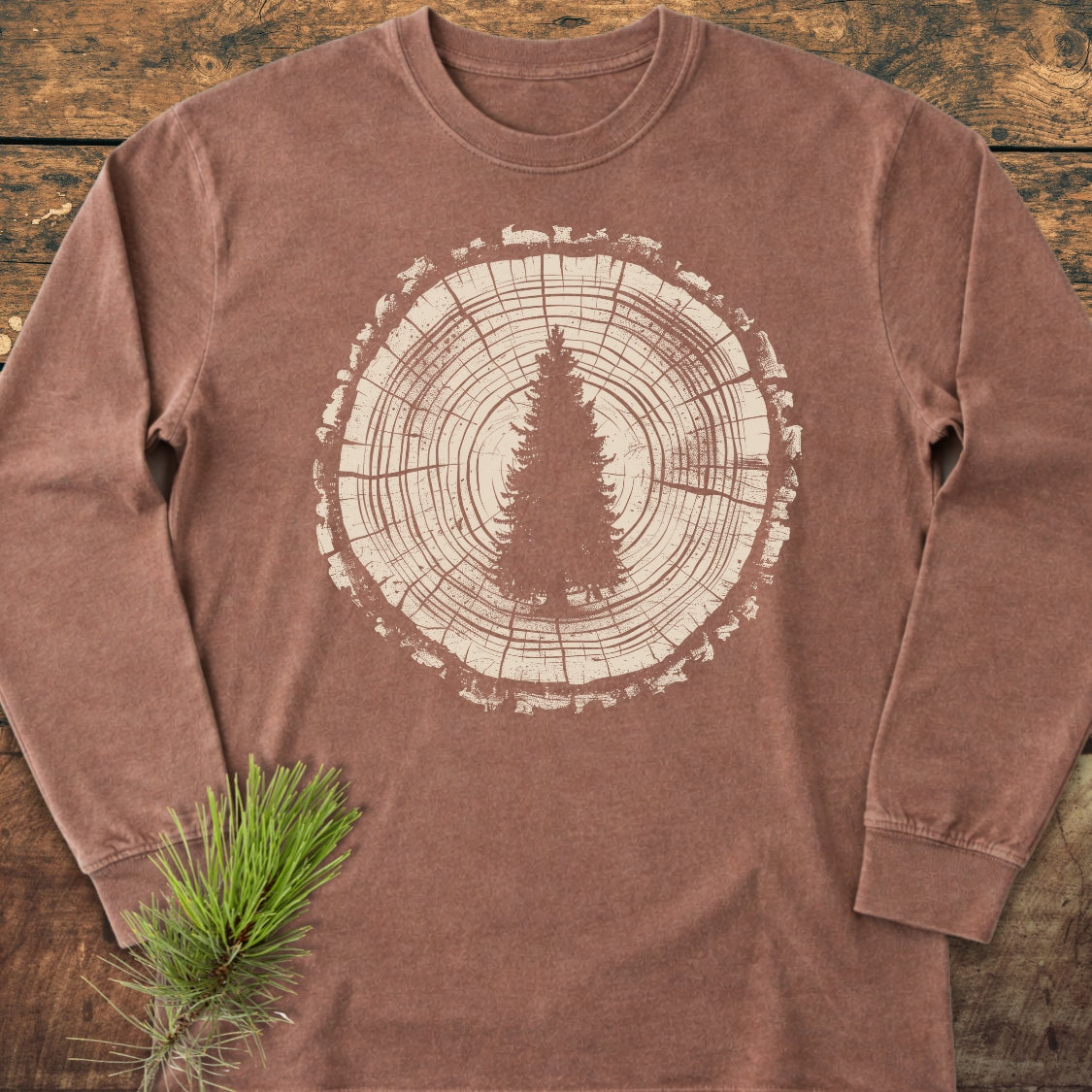 Rooted In The Wild Long-Sleeve Tee featuring a tree trunk graphic with an evergreen silhouette on soft, garment-dyed cotton, laid flat on a wooden surface with a pine sprig nearby.