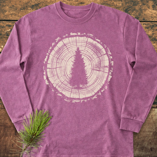 Rooted In The Wild Long-Sleeve Tee featuring a distressed tree trunk and evergreen graphic on a washed-out purple background, laid on wood, with a pine twig accentuating its forest-inspired design.
