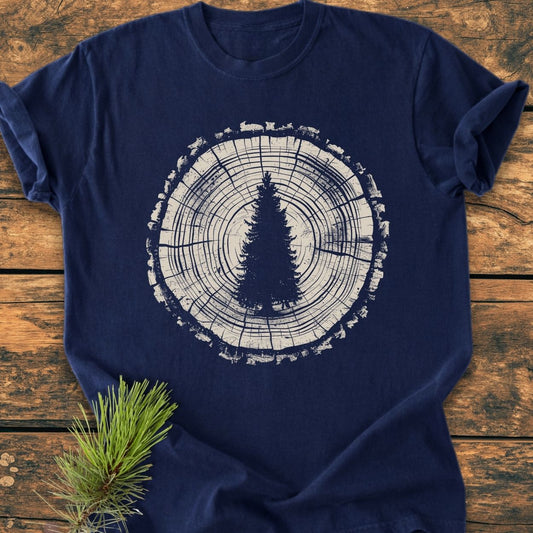 Rooted In The Wild - Vintage Wash Tee features a white tree-ring graphic with a pine tree silhouette on a navy-blue T-shirt, displayed on a wooden surface with a pine branch accent.