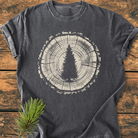 Rooted In The Wild - Vintage Wash Tee: Dark-gray T-shirt featuring a tree-ring graphic with an evergreen silhouette, laid on wood. Beside it is a real pine sprig, highlighting its nature-inspired design.