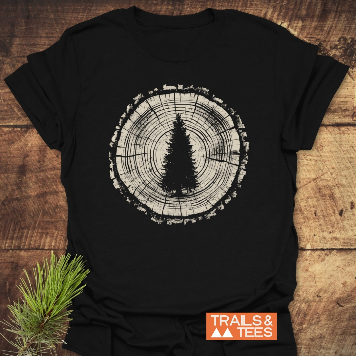 Rooted In Nature T-Shirt featuring a tree-ring and evergreen design on black fabric, laid on wood with a pine sprig. Includes “TRAILS & TEES” logo, emphasizing outdoor adventure.
