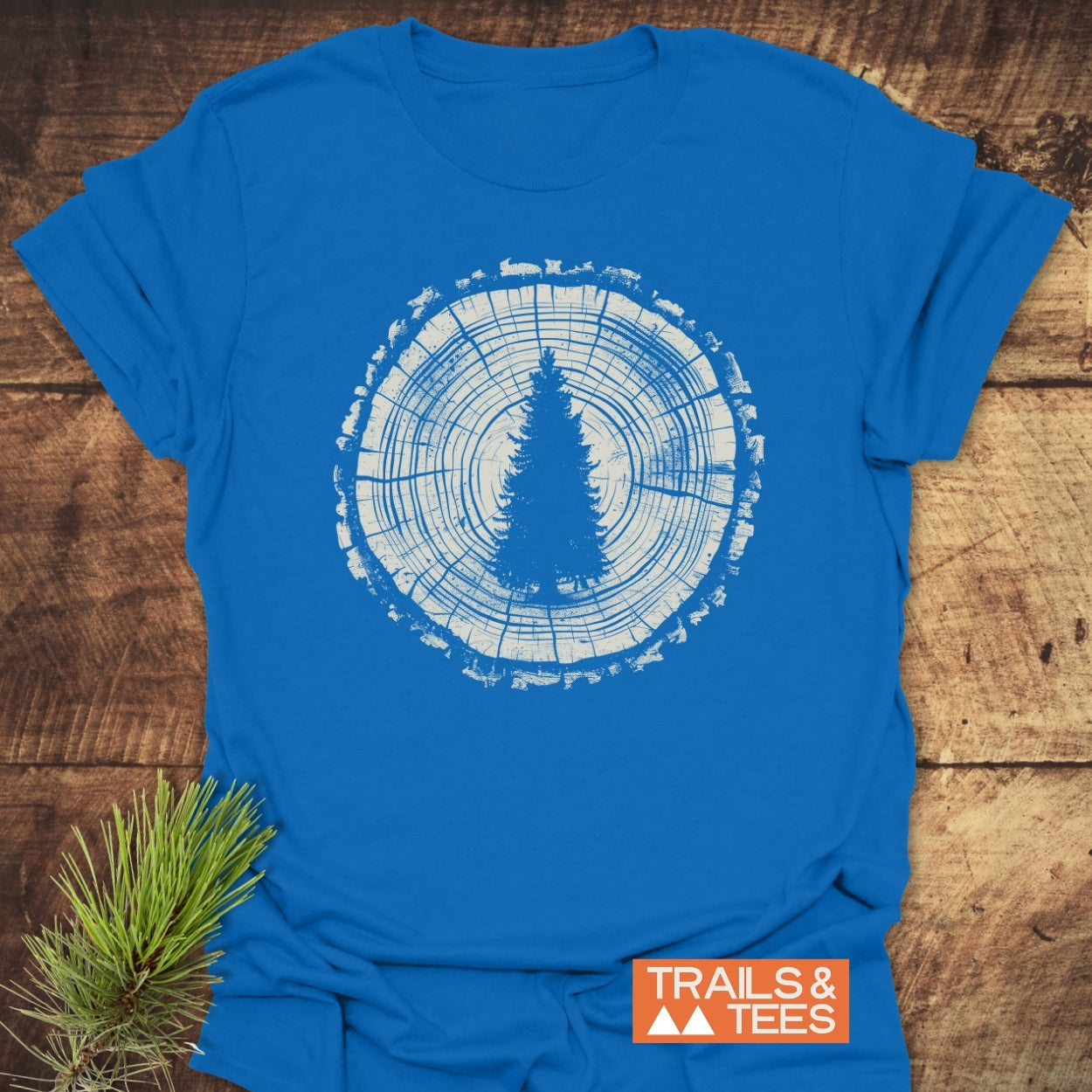 Rooted In Nature T-Shirt features a white tree-ring graphic with a pine silhouette, styled on a wooden surface with pine needles and the TRAILS & TEES logo in the corner.
