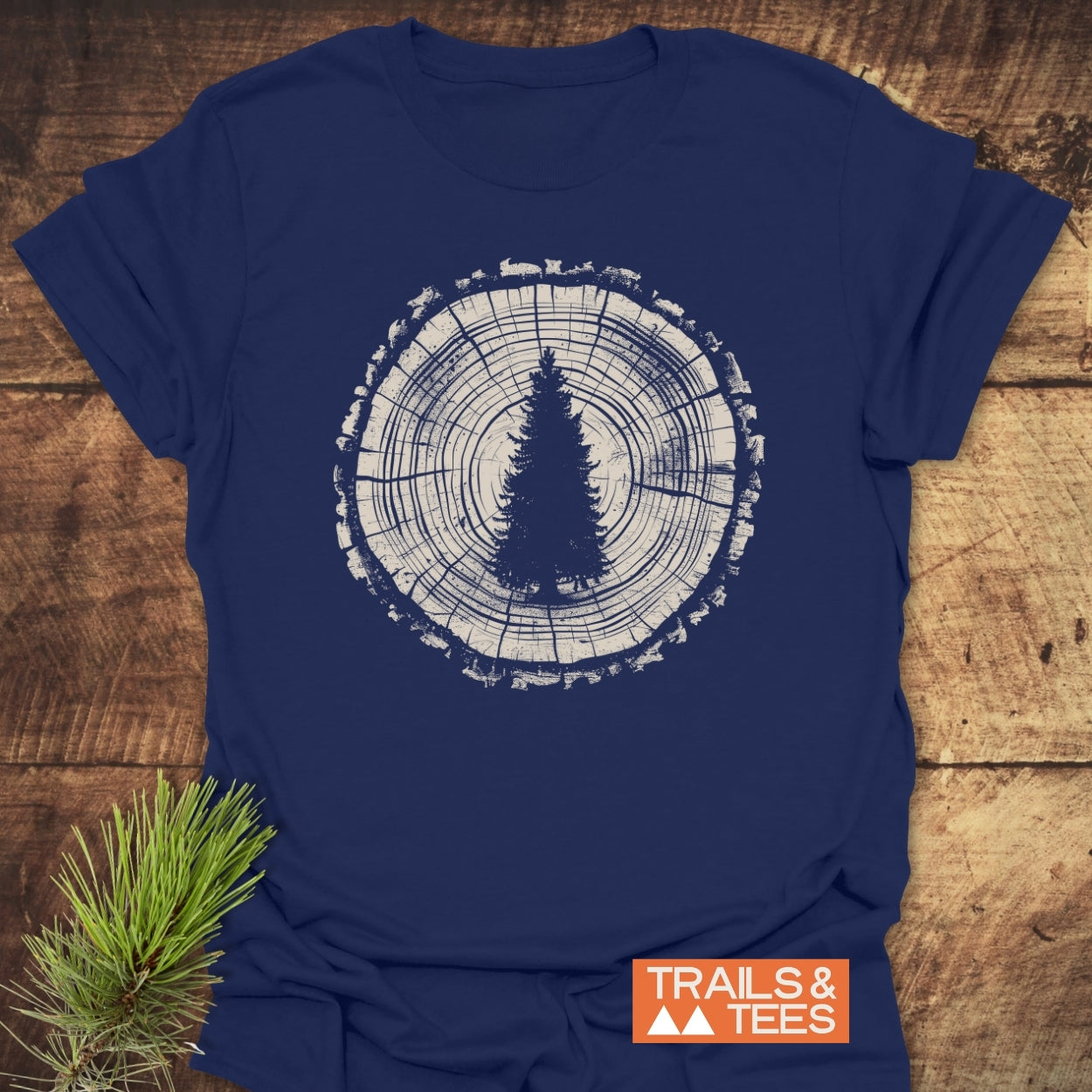 Rooted In Nature T-Shirt featuring a tree trunk graphic with an evergreen silhouette, laid on wood. Includes TRAILS & TEES logo and pine sprig, highlighting outdoor-inspired design and comfort.