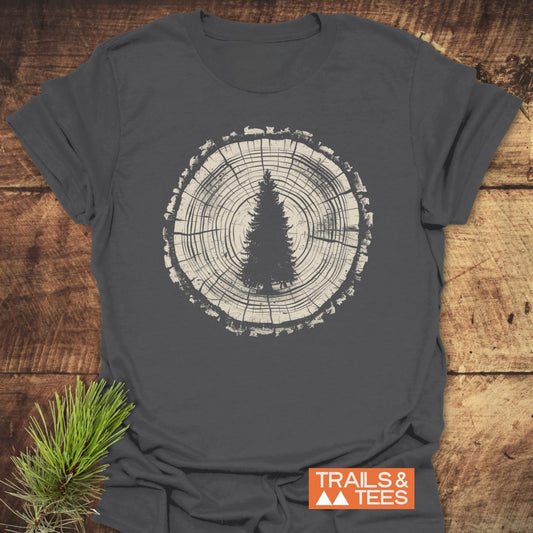 Rooted In Nature T-Shirt features a tree-ring cross-section with an evergreen silhouette, laid on wood. Includes a pine sprig and TRAILS & TEES logo, emphasizing outdoor adventure.