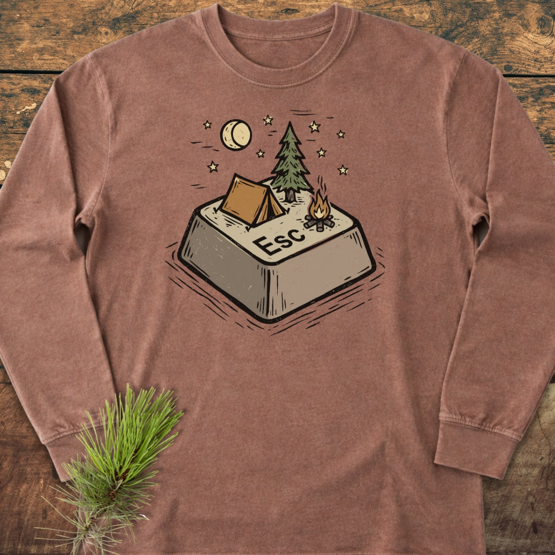 Escape Long-Sleeve Tee features a graphic of a campsite-themed Esc key with a tent, pine tree, and campfire, laid on wood with a pine sprig accentuating its cozy, adventure-inspired design.