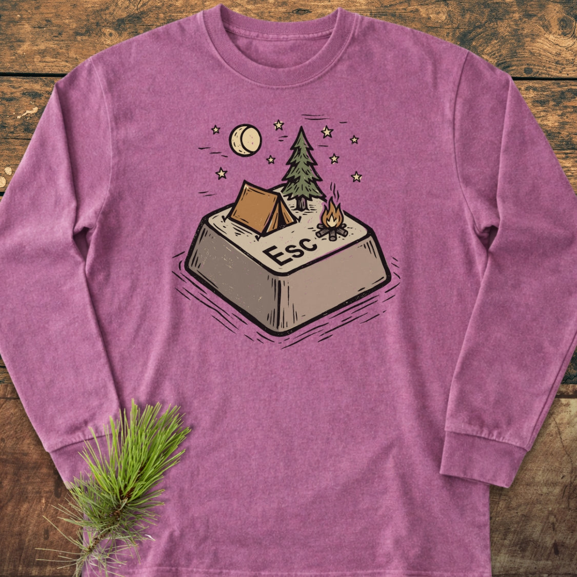 Escape Long-Sleeve Tee featuring an oversized Esc key with a campsite scene, laid flat on wood, accompanied by a pine sprig, highlighting its soft, garment-dyed 100% ring-spun cotton construction.