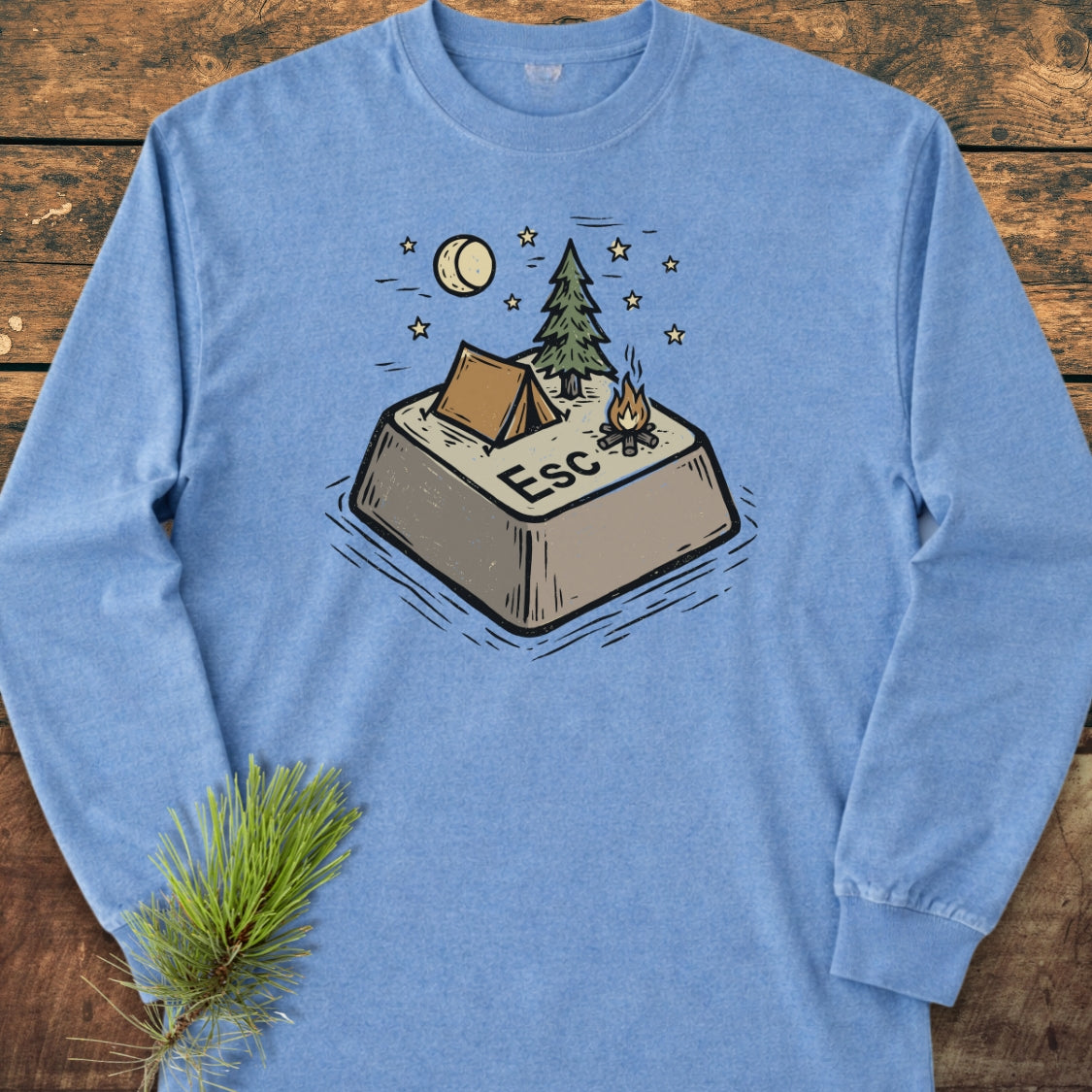 Escape Long-Sleeve Tee features a playful campsite illustration atop a giant Esc key. Laid flat on a wooden surface, this heather-blue shirt offers softness and style with its relaxed fit.