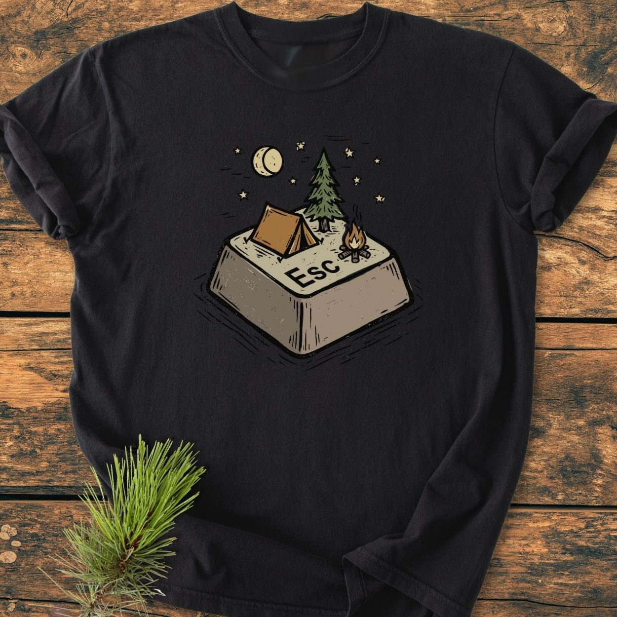 Escape - Vintage Wash Tee featuring a stylized Esc key as a campsite with a tent, pine tree, campfire, moon, and stars on a black T-shirt, laid on a wooden surface.