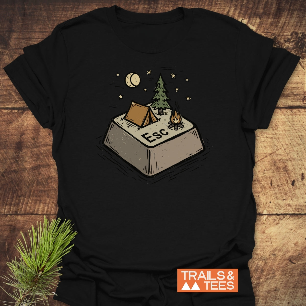 Escape T-Shirt featuring a stylized campsite graphic with a tent and campfire on a black cotton tee, laid on wood, next to a pine branch. Logo: TRAILS & TEES.