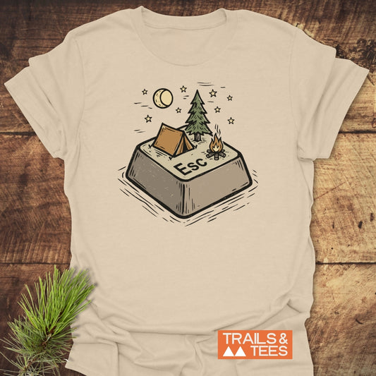 Escape T-Shirt featuring a whimsical campsite design with an Esc key, tent, pine tree, and campfire under stars and moon, complemented by the Trails & Tees logo on a beige backdrop.