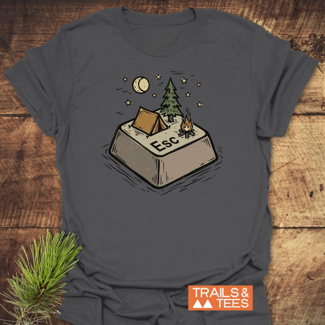 Escape T-Shirt featuring a campsite-themed Esc key illustration with a tent, fir tree, campfire, and moon. Crafted from soft cotton, ideal for outdoor enthusiasts. Includes TRAILS & TEES logo.