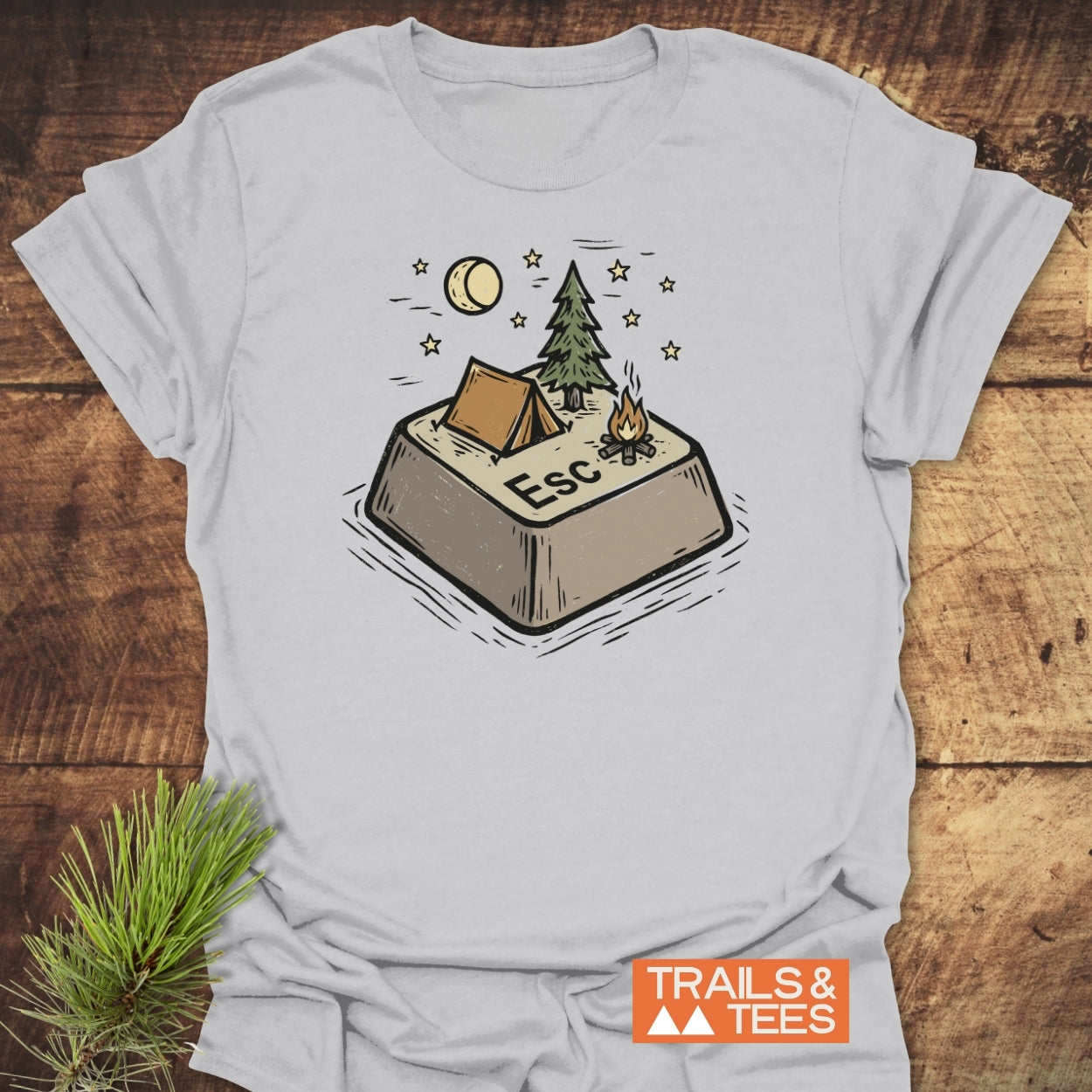 Escape T-Shirt featuring a playful campsite illustration on a giant Esc key, with a tent, pine tree, campfire, moon, and stars. Trails & Tees branding visible.