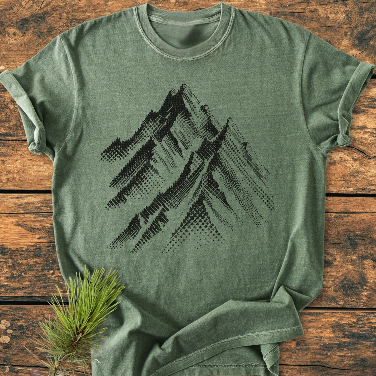 Take Me Higher - Vintage Wash Tee features a stylized mountain-range design on a soft green, rolled-sleeve T-shirt, laid on wood, complemented by a pine sprig for an outdoor feel.