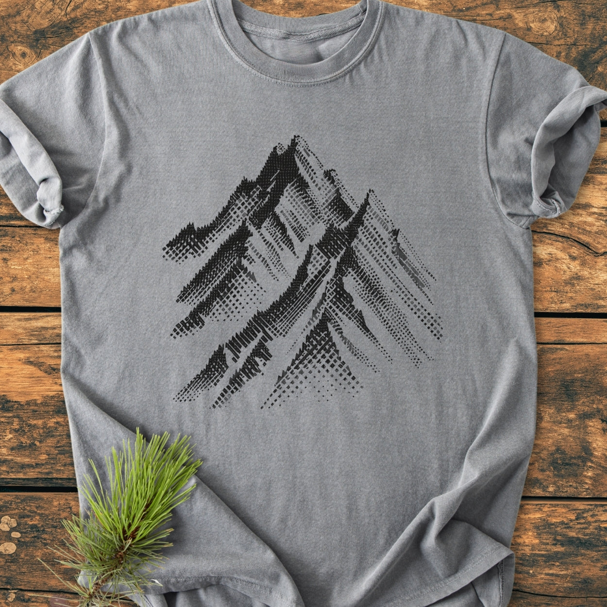 Take Me Higher - Vintage Wash Tee features a heather-gray color with a halftone mountain range design, laid flat on wood. A sprig of pine needles accents the scene.