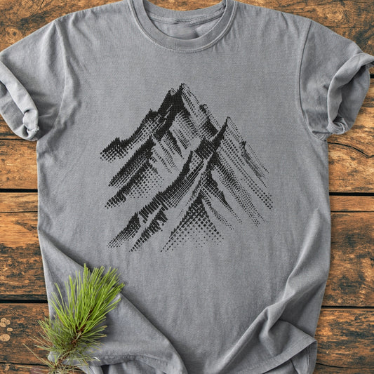Take Me Higher - Vintage Wash Tee features a heather-gray color with a halftone mountain range design, laid flat on wood. A sprig of pine needles accents the scene.