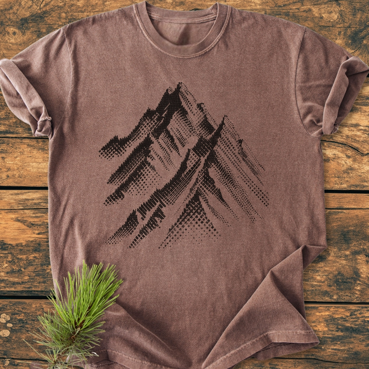 Take Me Higher - Vintage Wash Tee features a stylized mountain silhouette on a soft-washed, earth-tone T-shirt laid flat on wood, with a pine sprig accentuating its rustic charm.