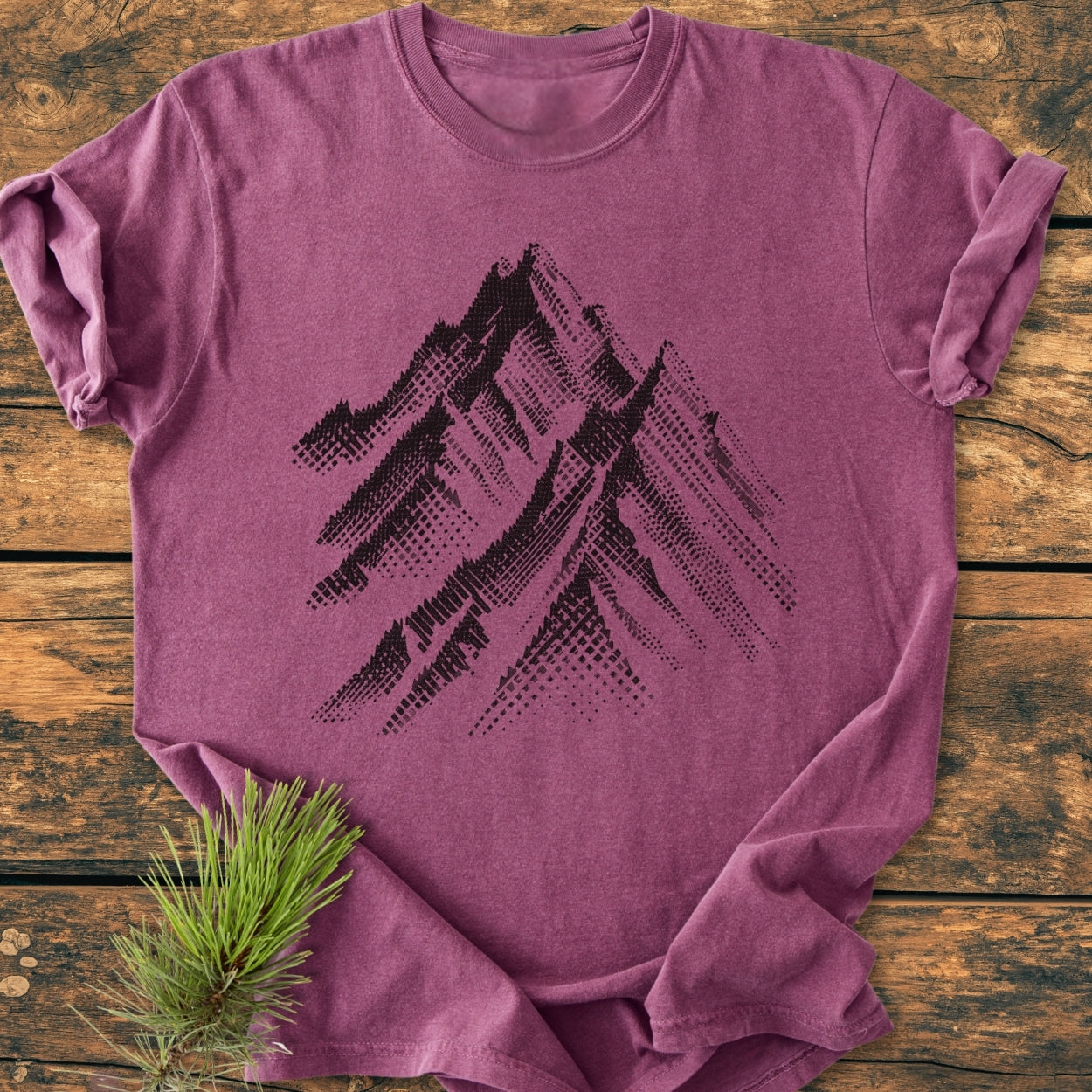 Take Me Higher - Vintage Wash Tee features a heather-purple tone with black mountain peaks graphic. Displayed on wood, pine sprig beside, emphasizing a relaxed, lifestyle vibe.