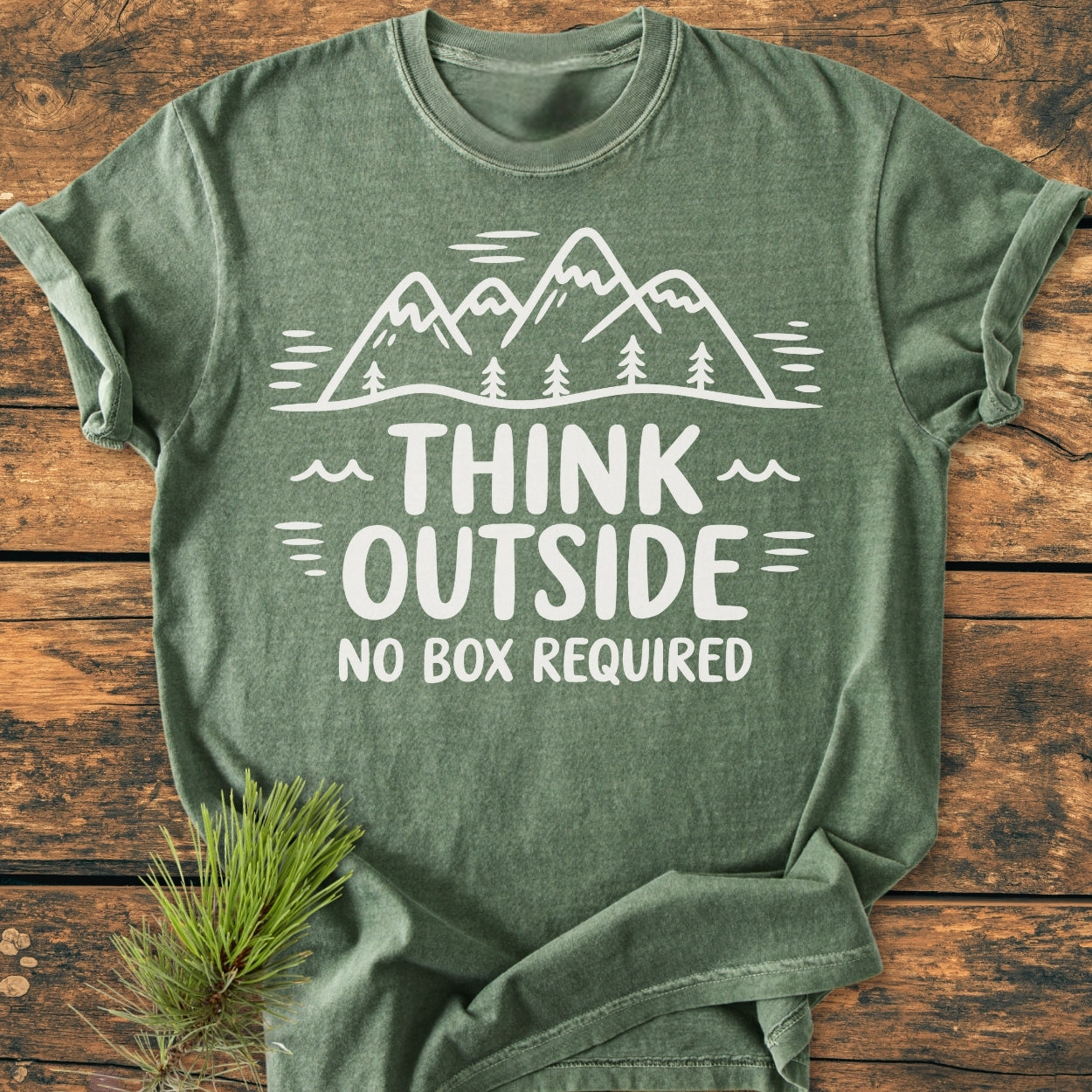 Think Outside - Vintage Wash Tee in heather green, features a white line-drawing of mountains and pine trees with the slogan THINK OUTSIDE NO BOX REQUIRED, laid on a wooden surface with an evergreen sprig.