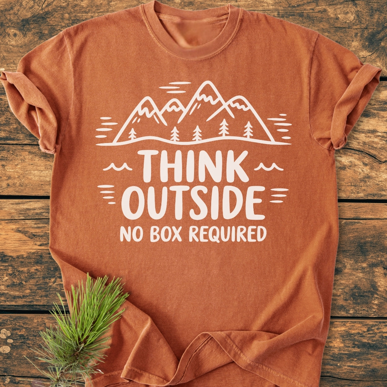 Think Outside - Vintage Wash Tee features a rust-orange hue with a white line-drawing of mountains and trees, paired with the slogan THINK OUTSIDE NO BOX REQUIRED, displayed on a wooden surface with a pine branch.