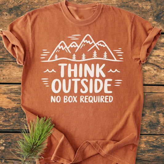 Think Outside - Vintage Wash Tee features a rust-orange hue with a white line-drawing of mountains and trees, paired with the slogan THINK OUTSIDE NO BOX REQUIRED, displayed on a wooden surface with a pine branch.