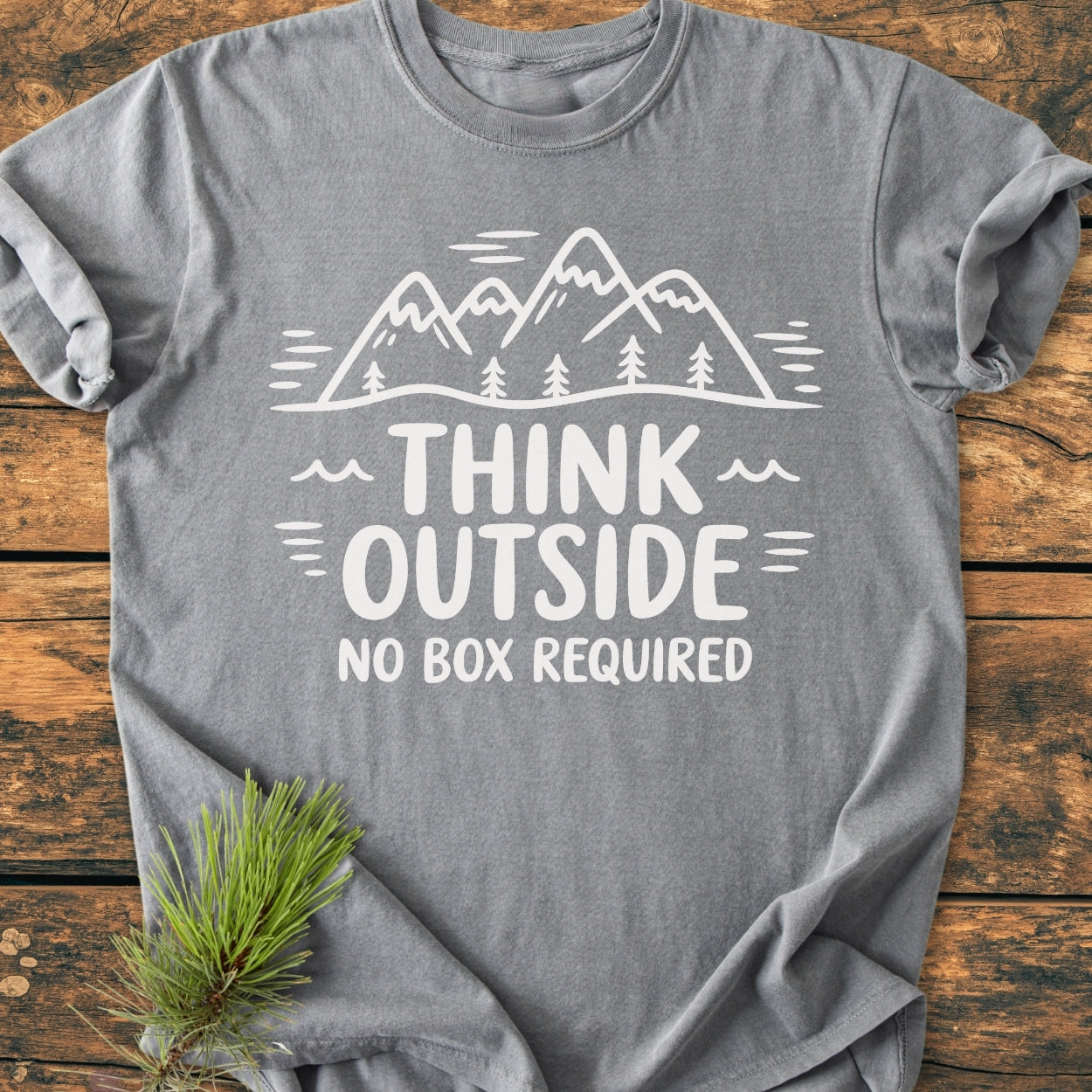 Think Outside - Vintage Wash Tee: Heather-gray T-shirt with mountain-and-pine-tree graphic and THINK OUTSIDE, NO BOX REQUIRED text, styled on a wooden surface with a pine branch beside it.