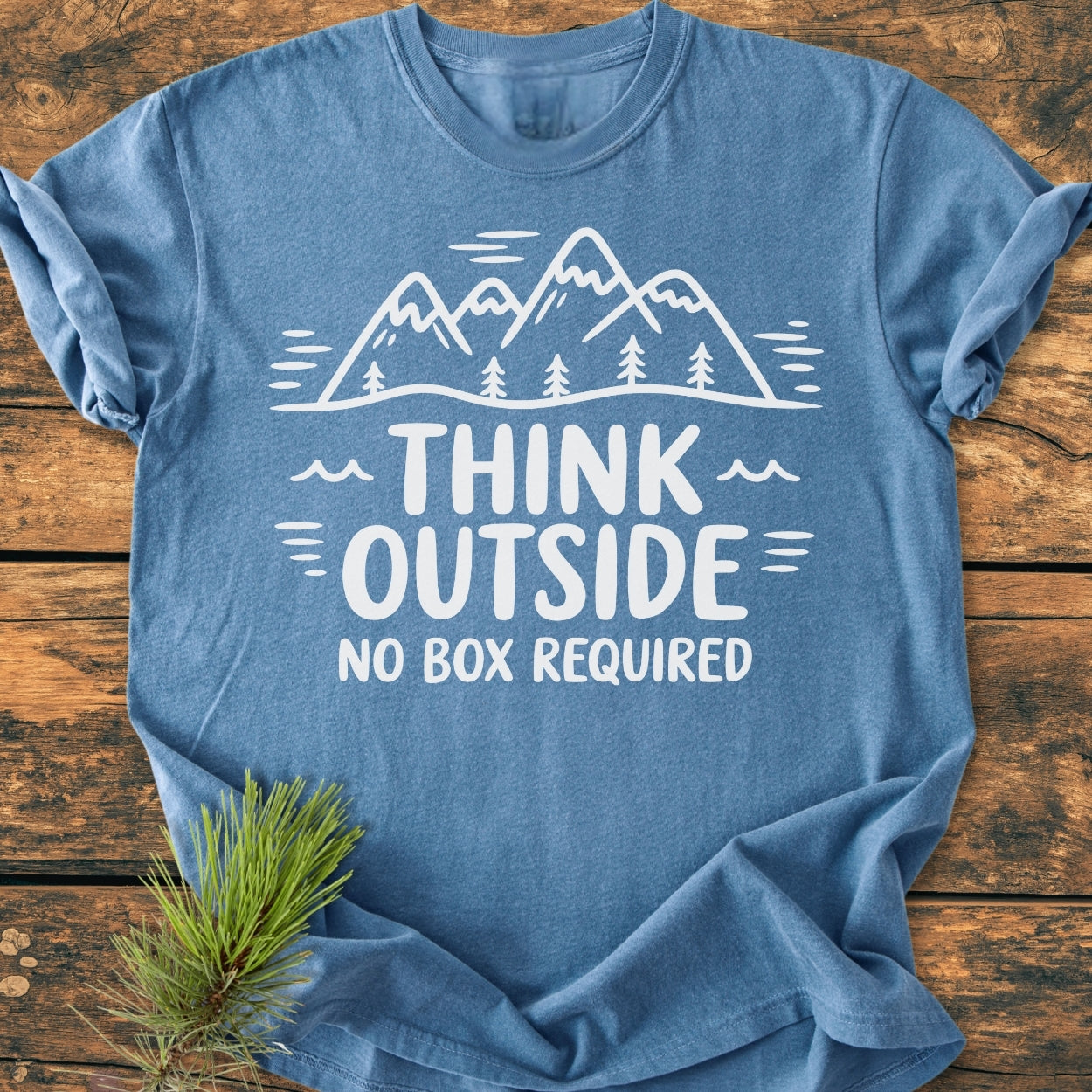 Think Outside - Vintage Wash Tee features a soft-blue color with a white mountain line-drawing and slogan, displayed on rustic wood, complemented by an evergreen sprig, highlighting its outdoor theme.