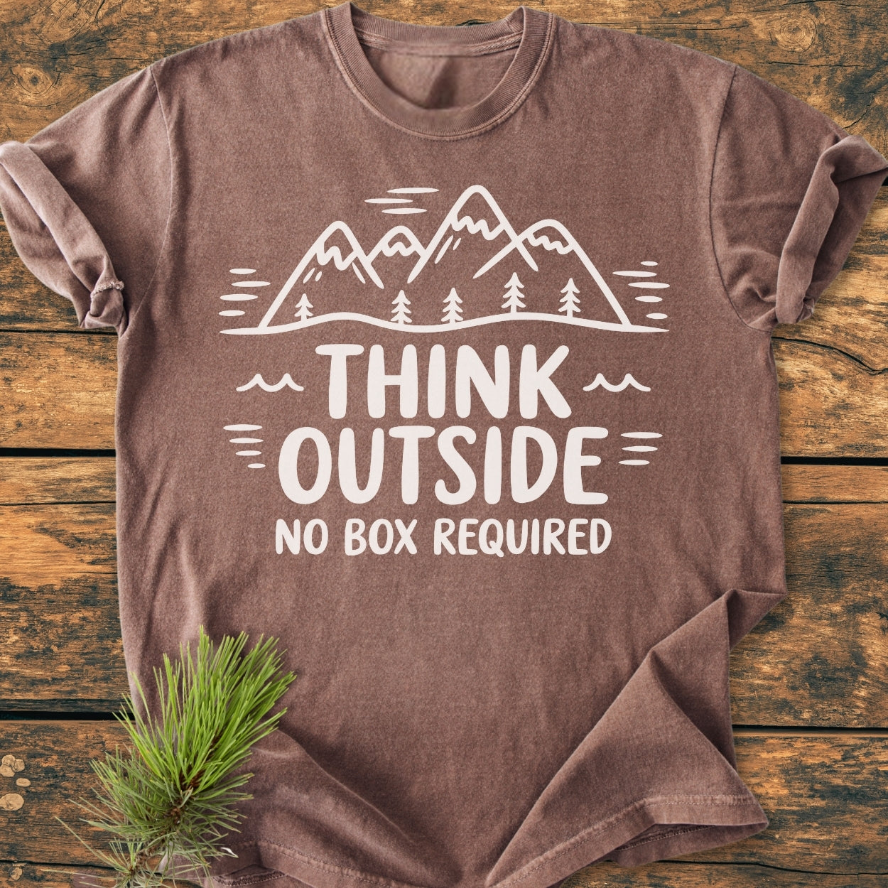 Think Outside - Vintage Wash Tee: Brown T-shirt with white line-drawing of mountains and trees, slogan THINK OUTSIDE NO BOX REQUIRED, on a rustic wooden surface with pine sprig accent.