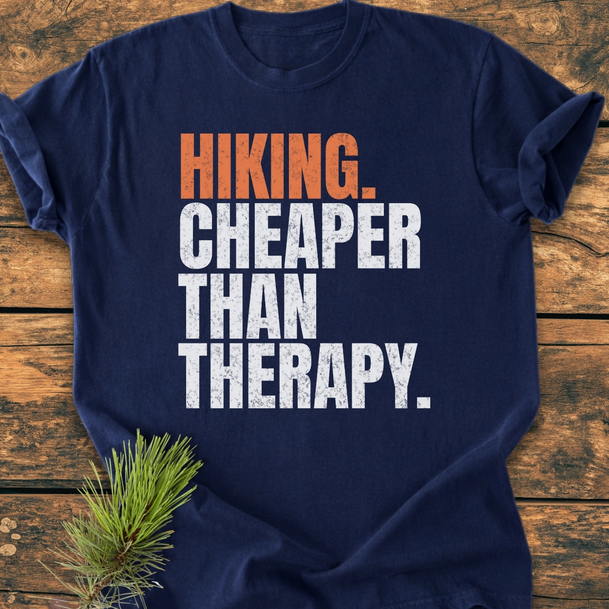 Navy Hiking Cheaper Than Therapy - Vintage Wash Tee on wood, featuring orange and white slogan, with pine sprig accentuating its rustic, outdoorsy appeal. Crafted from soft, ring-spun cotton for comfort.