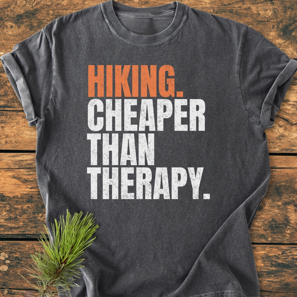 Hiking Cheaper Than Therapy - Vintage Wash Tee featuring bold block letters on a charcoal-gray T-shirt, laid flat on wood, with a pine sprig accentuating the cozy, relaxed design.