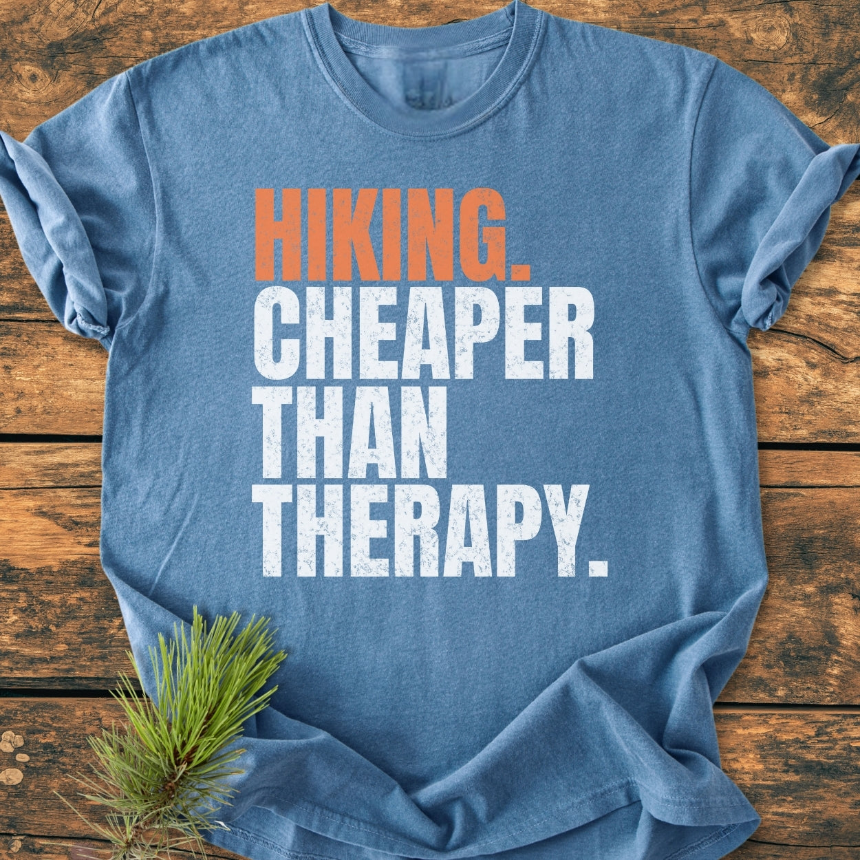 Hiking Cheaper Than Therapy - Vintage Wash Tee displayed on wood, features bold block text, a pine sprig accent, and a soft, durable, relaxed fit, made from pre-shrunk, 100% ring-spun cotton.
