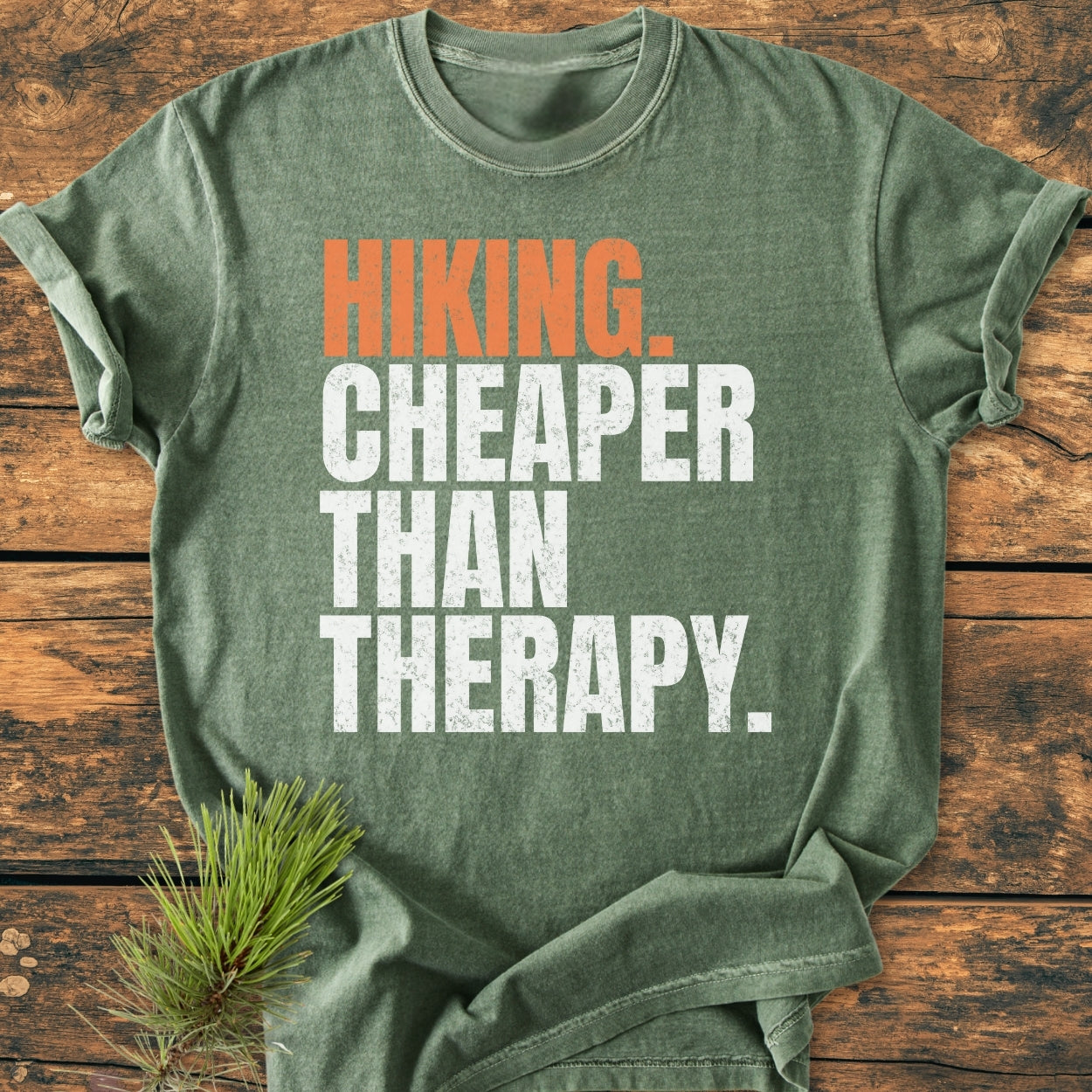 Heather-green Hiking Cheaper Than Therapy - Vintage Wash Tee laid flat on wood, featuring bold slogan. Includes pine sprig accentuating the outdoors theme. Garment-dyed, soft-washed, relaxed fit, durable stitching.