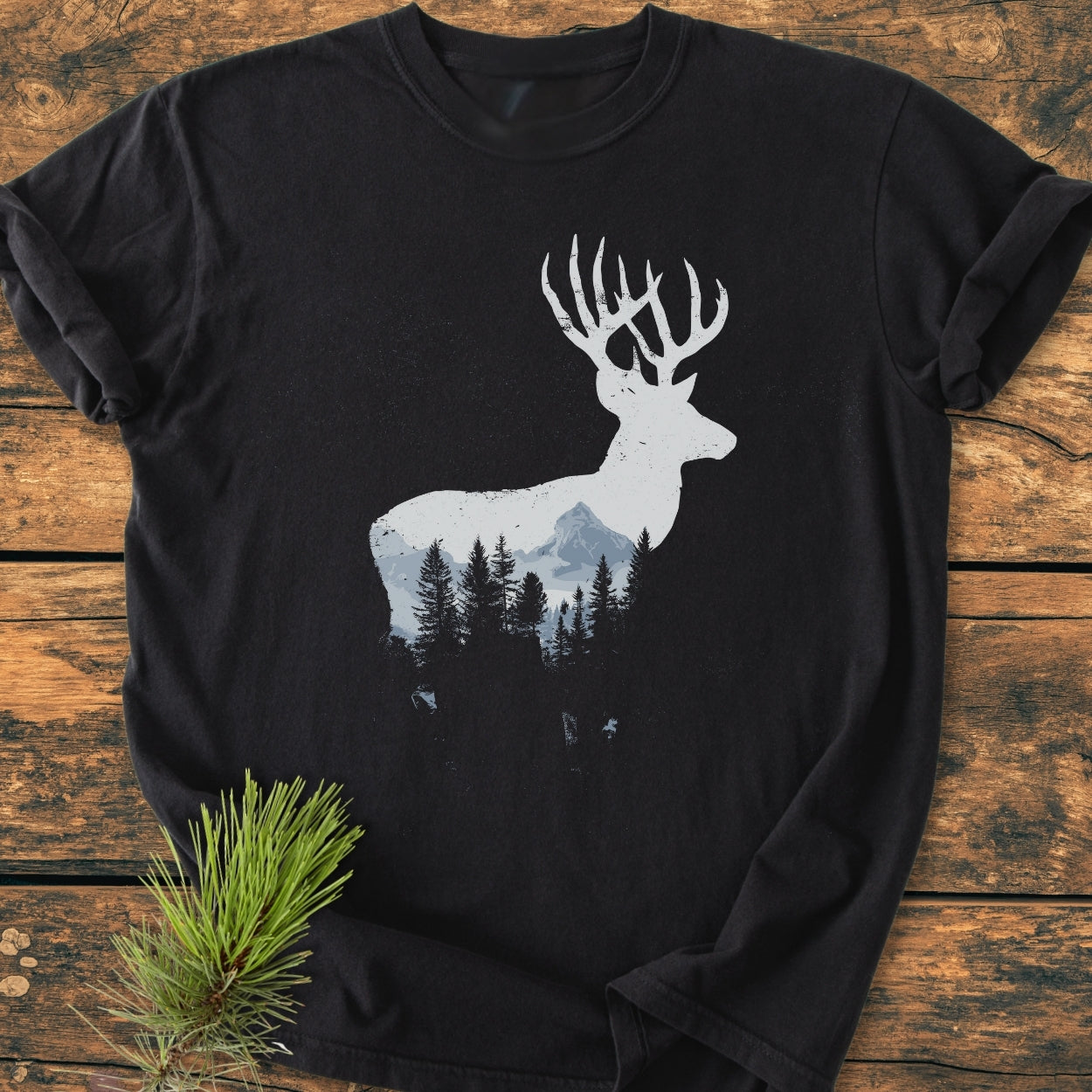 Deer Silhouette - Vintage Wash Tee: Black T-shirt with a white stag silhouette featuring a mountain and pine design, laid on wood. Includes a pine sprig, emphasizing the woodland theme.
