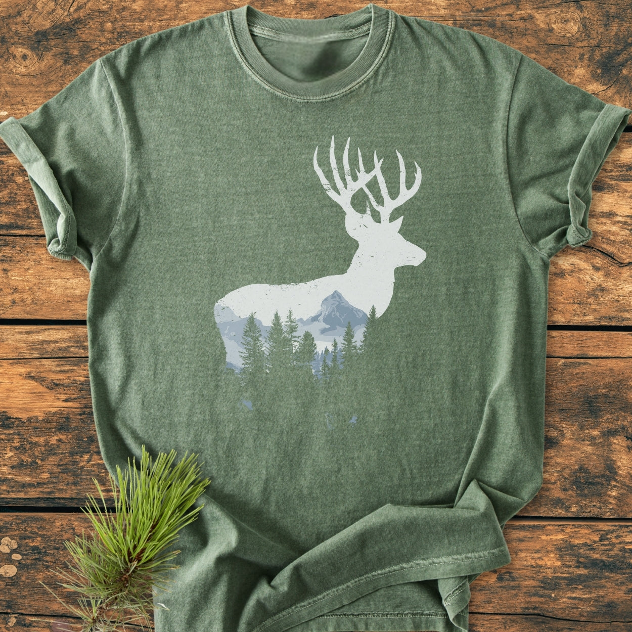 Deer Silhouette - Vintage Wash Tee: Green heathered short-sleeve T-shirt with a white stag silhouette featuring a forest-and-mountain graphic, laid on a wooden surface with a pine sprig for styling.