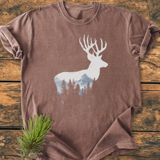 Deer Silhouette - Vintage Wash Tee features a brown, short-sleeve T-shirt with a white stag silhouette showcasing mountains and pine trees, laid flat on a wooden surface with a decorative pine sprig.